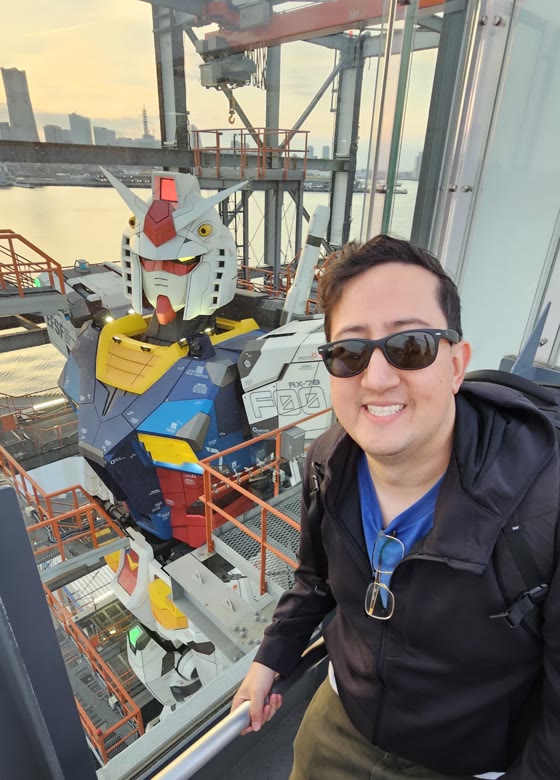 Chris in sunglasses beside a life-size RX-78 Gundam statue, harbour and sunset behind it.